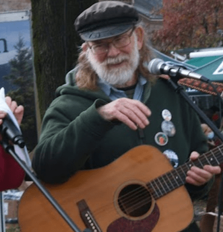 color photo of a white man with a white beard and a guitar. He's wearing a green hat and green coat
