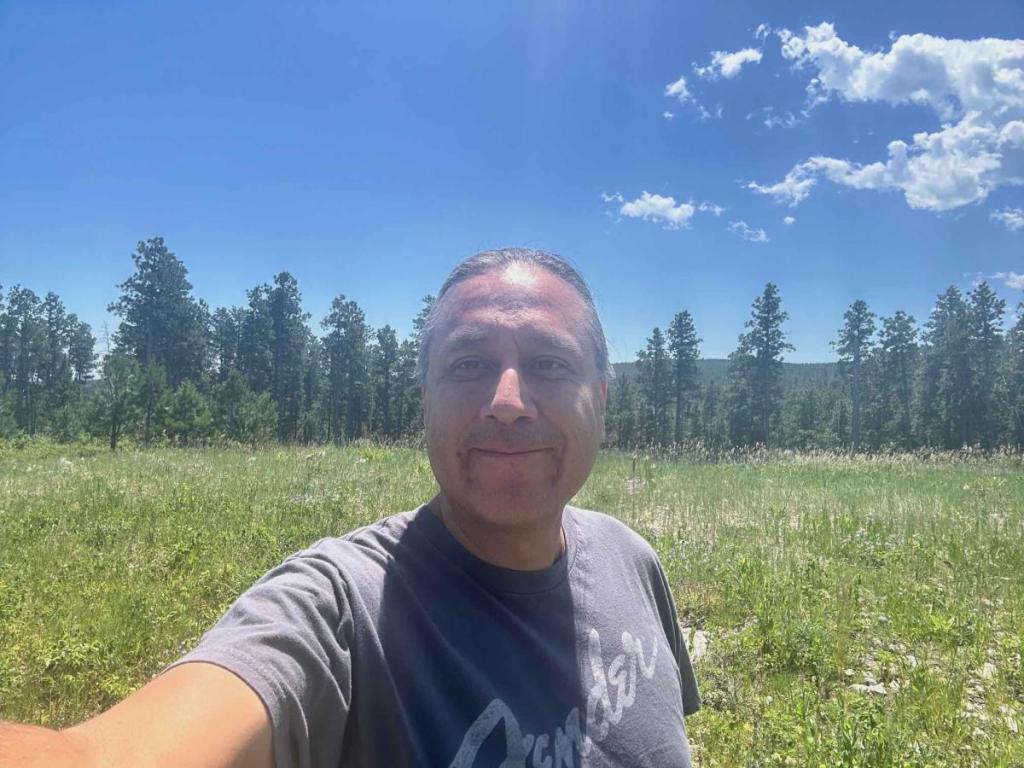 selfie of a smiling man wearing a Fender tshirt, hair pulled back. photo taken in prairie with woods in background