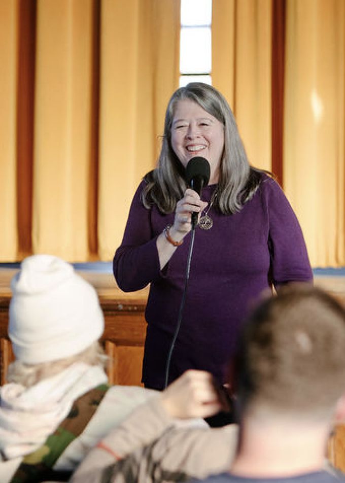 photo of a woman smiling and holding a microphone. She's wearing a purple dress, long gray hair not tied up, and she's speaking to a crowd