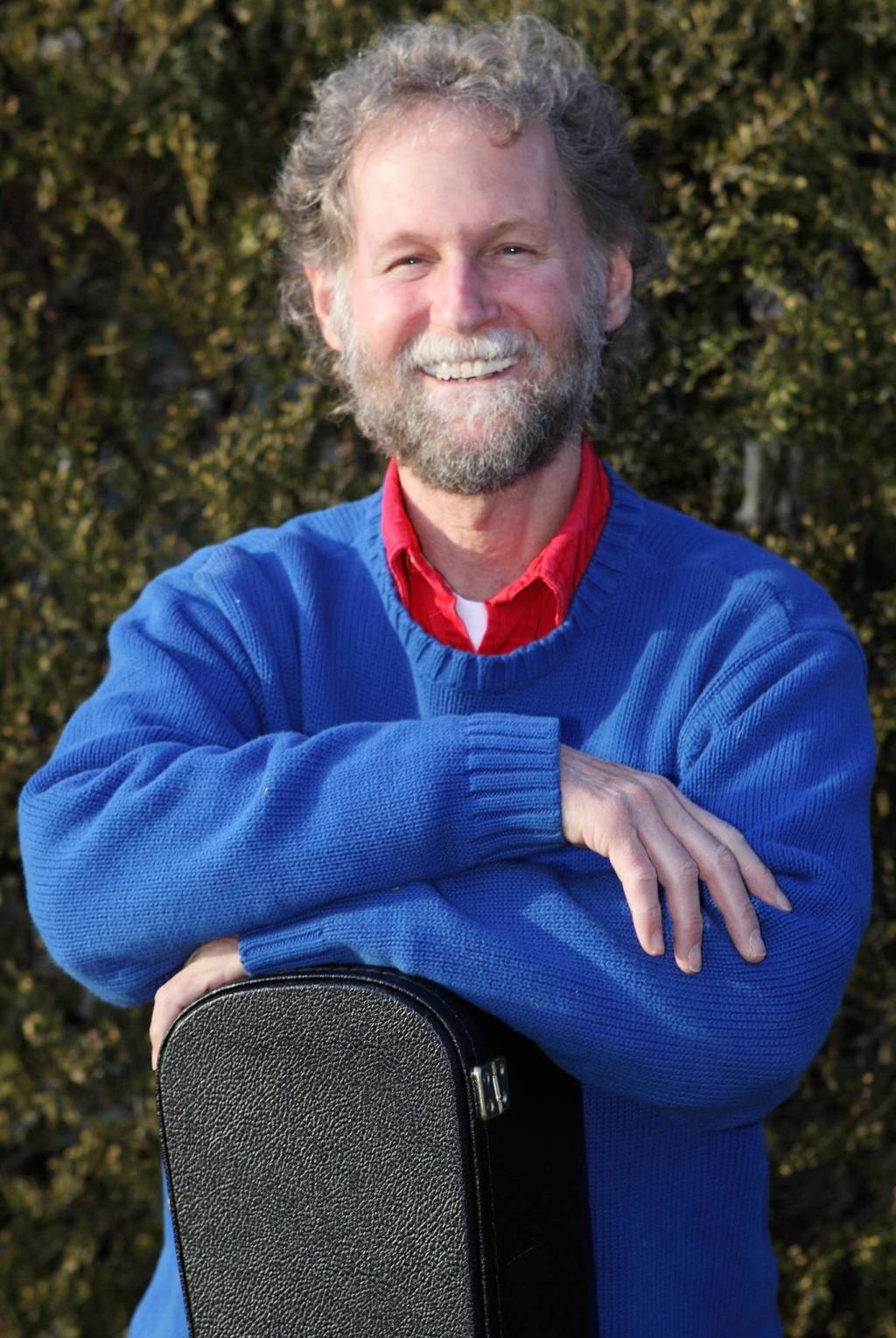 smiling man with arms crossed over a guitar case. he's wearing a blue sweater and it outdoors